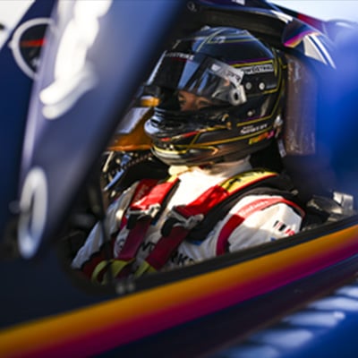 Race car driver wearing helmet and harness, seated inside a cockpit with colorful trim