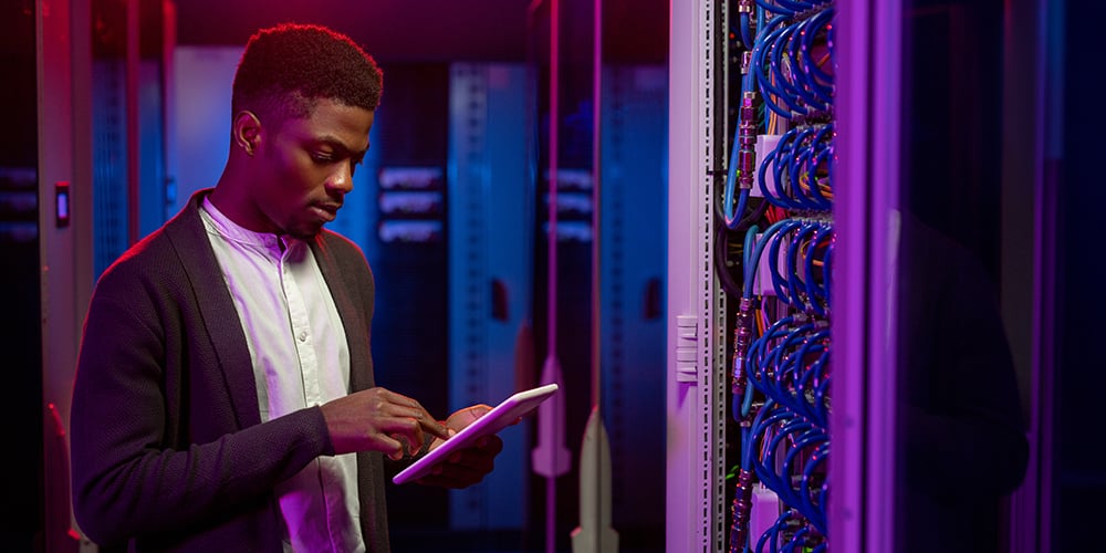 Person using a tablet in a server room, standing beside a rack of network cabling under colored lighting