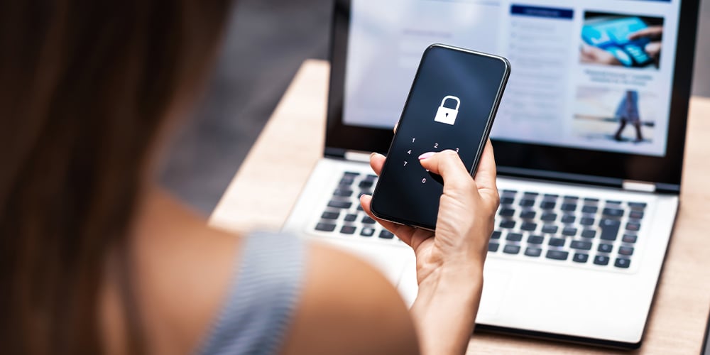 Hand holding a smartphone with a lock icon, with a laptop in the background showing web content