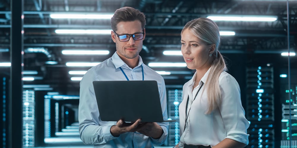 Two people reviewing a laptop in a data centre, surrounded by server racks and cool blue lighting