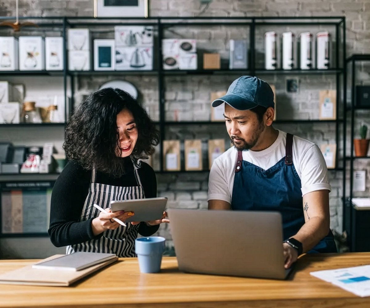 Two people working at a counter, one holding a tablet while the other uses a laptop, with products displayed on shelves behind