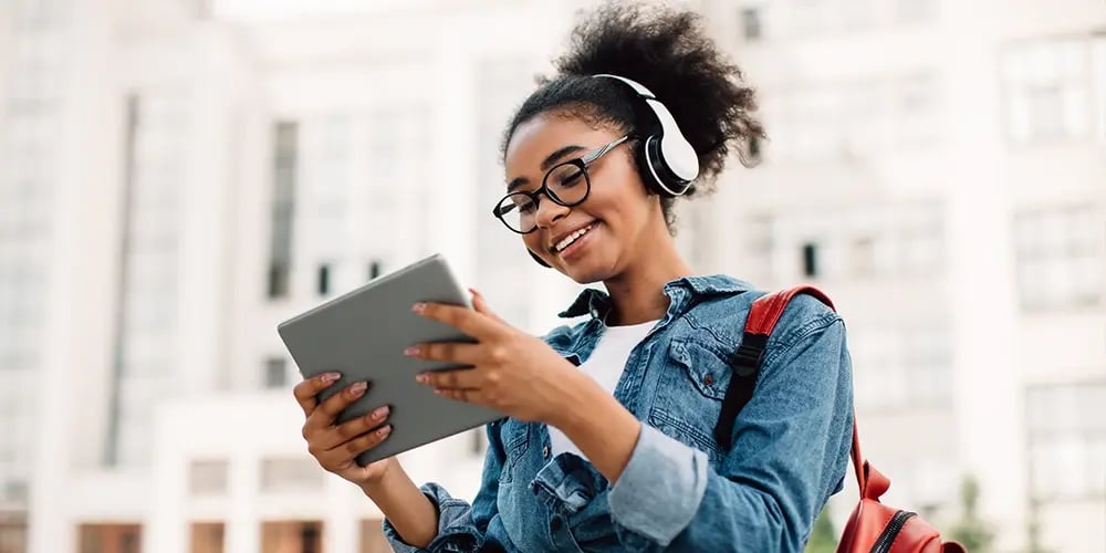Person wearing headphones and carrying a red backpack while holding a tablet outdoors.