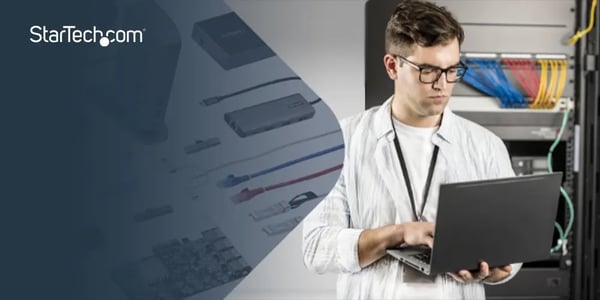 Person working on a laptop near a server rack, with various tech accessories displayed in the background.
