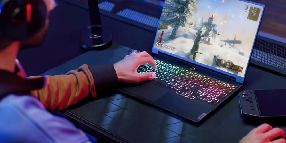 Gaming laptop with a colorful RGB backlit keyboard displaying a snowy action game on the screen, placed on a desk with a game controller nearby.
