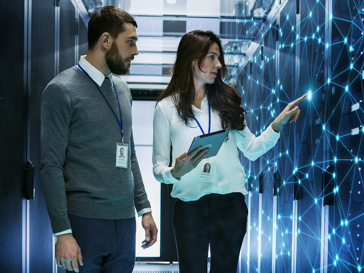 Two people in a server room reviewing data on a tablet and pointing at a network panel.