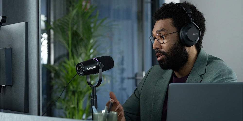 Person speaking into a Shure microphone while wearing headphones at a desk.