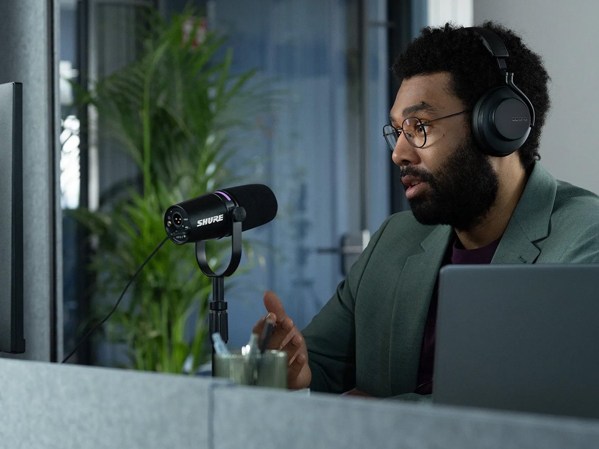 Person speaking into a Shure microphone while wearing headphones at a desk.
