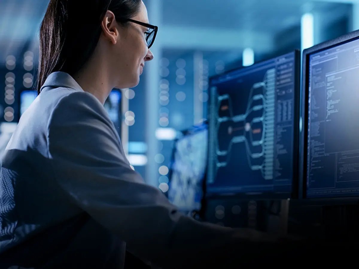 A person in glasses works at multiple monitors displaying complex data and graphs in a dimly lit, high-tech office, conveying a focused, analytical atmosphere.