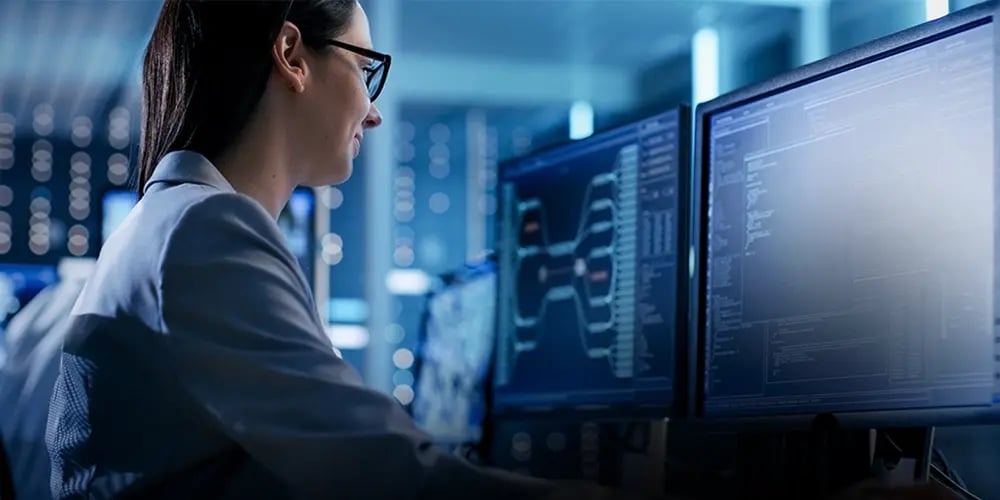 A person in glasses works at multiple monitors displaying complex data and graphs in a dimly lit, high-tech office, conveying a focused, analytical atmosphere.
