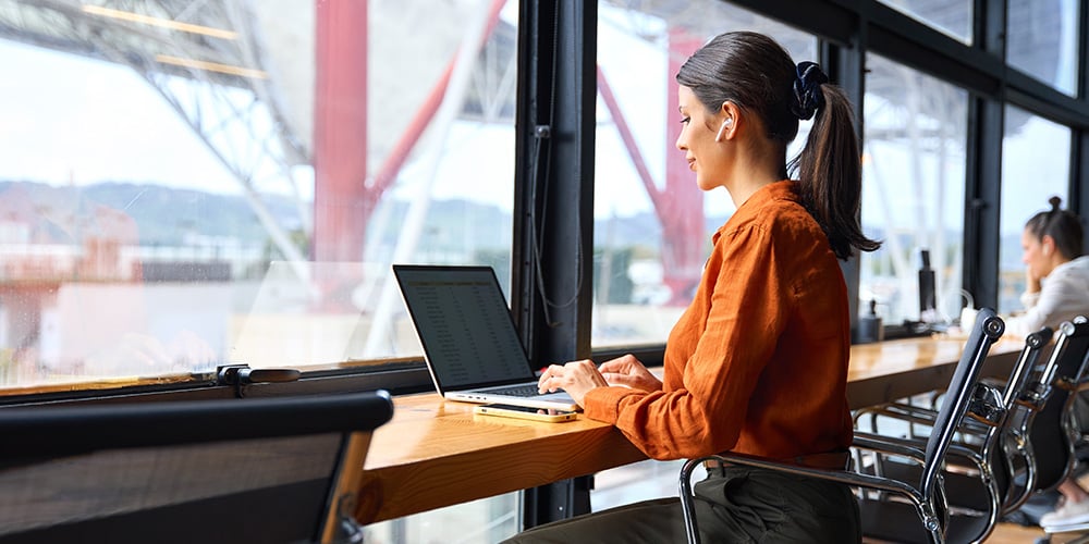 Person working on a laptop at a desk beside a large window in a modern office setting.