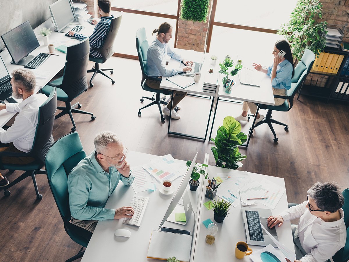 Open‑plan office with people working at desks, using computers and paperwork in a collaborative environment.