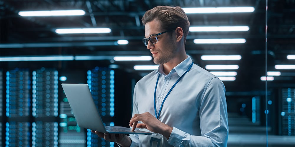 Technician standing in a data centre using a laptop to monitor server infrastructure.