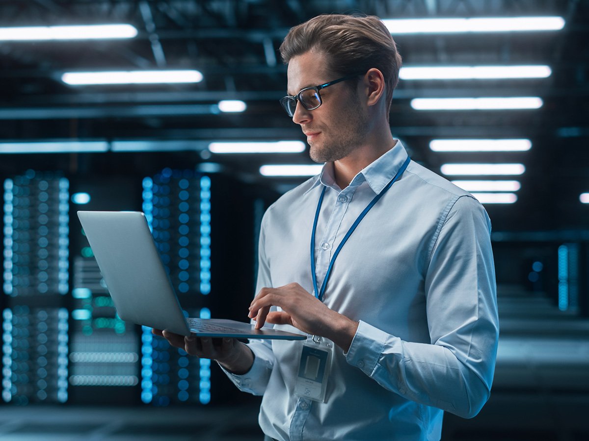Technician standing in a data centre using a laptop to monitor server infrastructure.