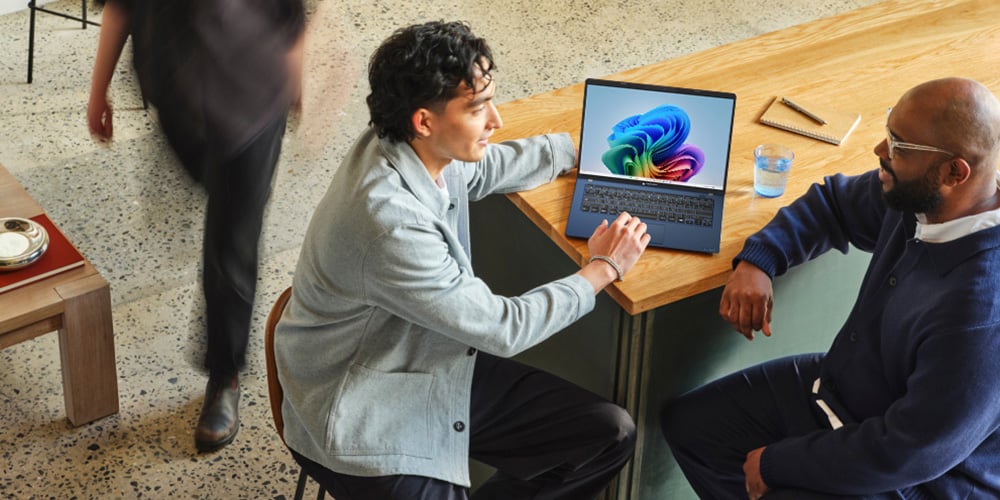 Two people sitting at a wooden table with a laptop displaying a colorful screen.