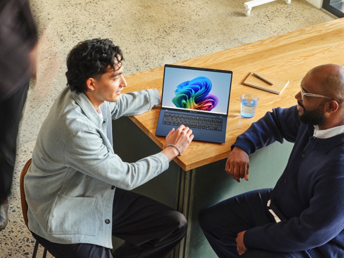 Two people sitting at a wooden table with a laptop displaying a colorful screen.