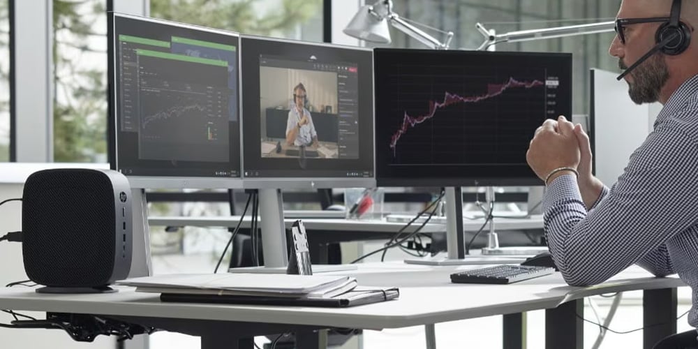 Person at a desk with three monitors showing charts, graphs, and a video call.