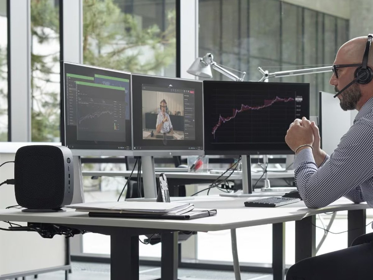 Person at a desk with three monitors showing charts, graphs, and a video call.