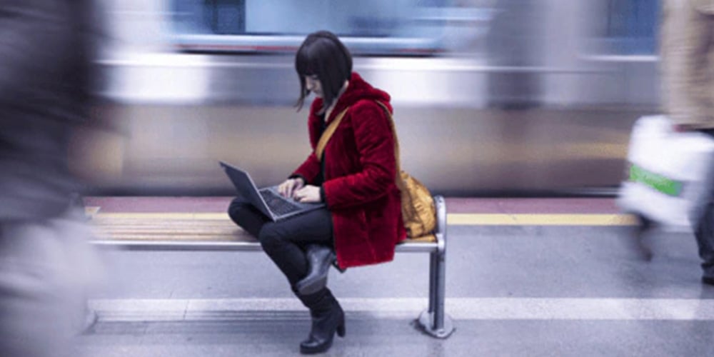 Individual using a laptop while seated on a bench at a train station.