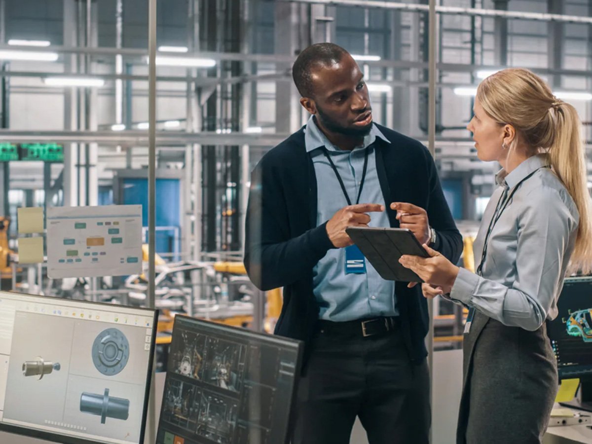 Two people standing in a factory office reviewing data on a tablet with monitors nearby.
