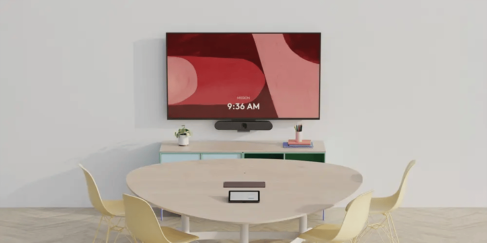 Minimalist conference room with a round table and four chairs. A large screen displays abstract art and the time, 9:36 AM. Calm and modern ambiance.