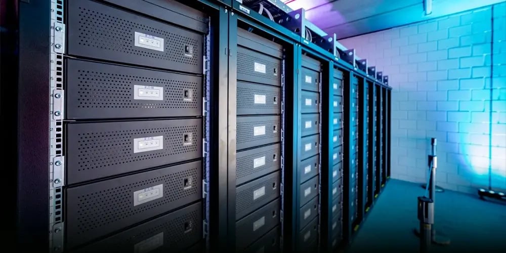 A row of black server racks in a data center with blue and purple ambient lighting, creating a modern and high-tech atmosphere.