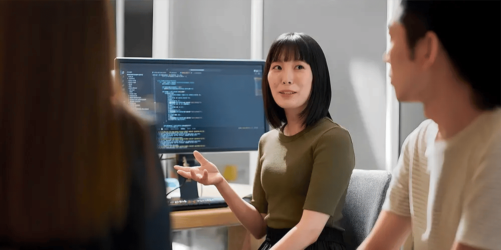 A woman in a green shirt gestures while discussing something with two others. Behind her is a computer screen displaying code. The atmosphere is collaborative.