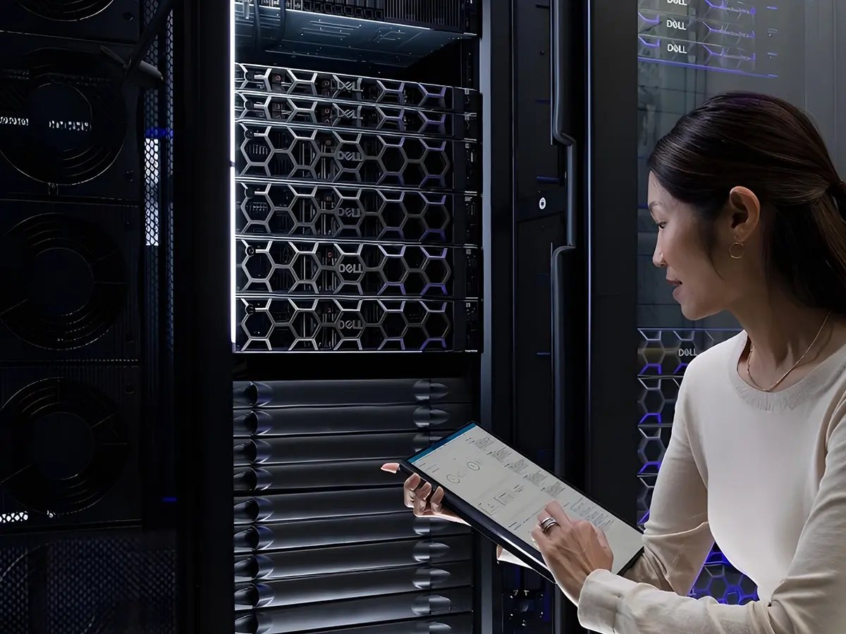 A woman examines a server rack while holding a tablet. The room is dimly lit, highlighting the sleek, modern technology. The tone is professional and focused.
