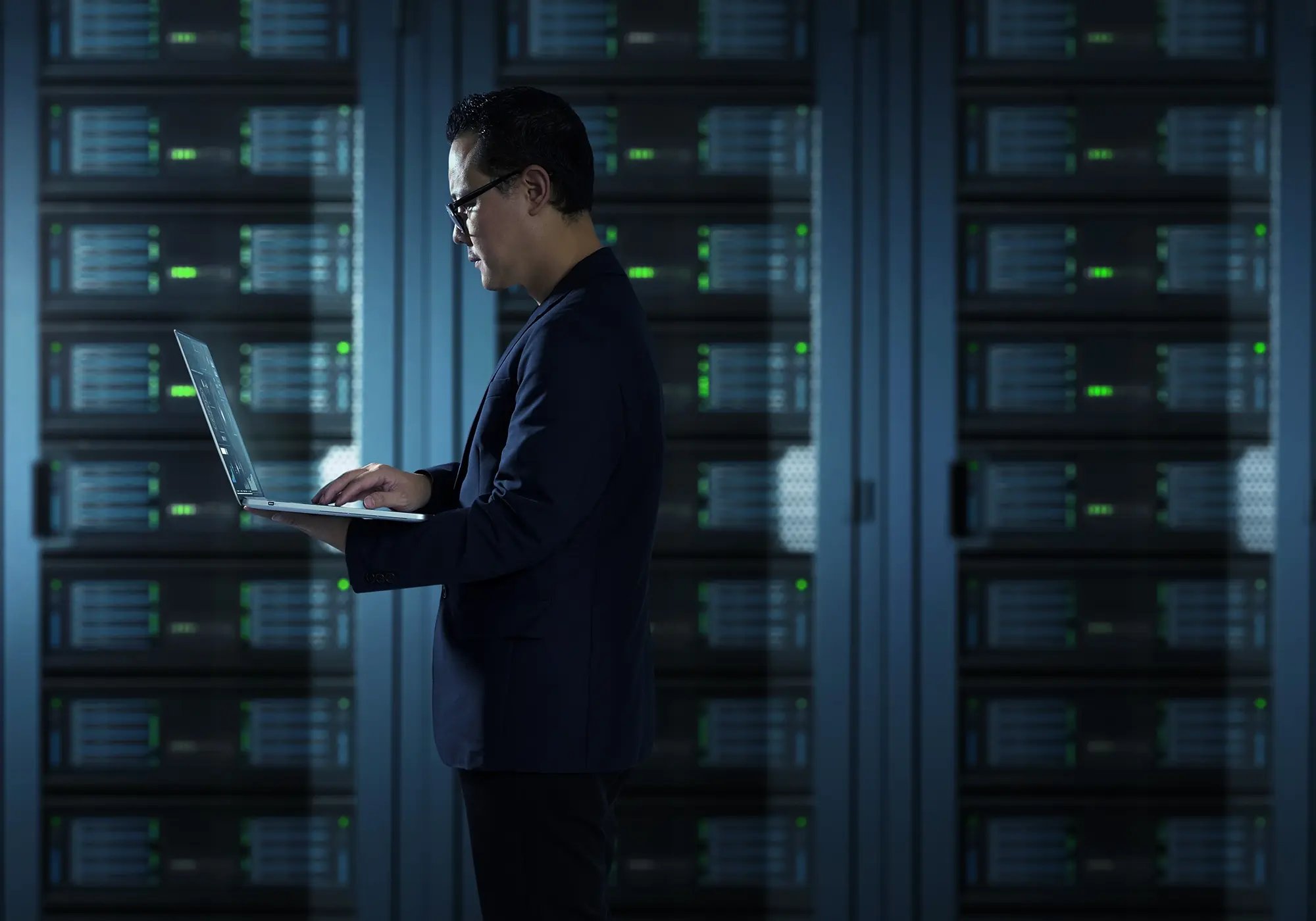 Person standing in a server room holding a laptop, surrounded by rows of server racks with green indicator lights, representing secure data erasure processes.