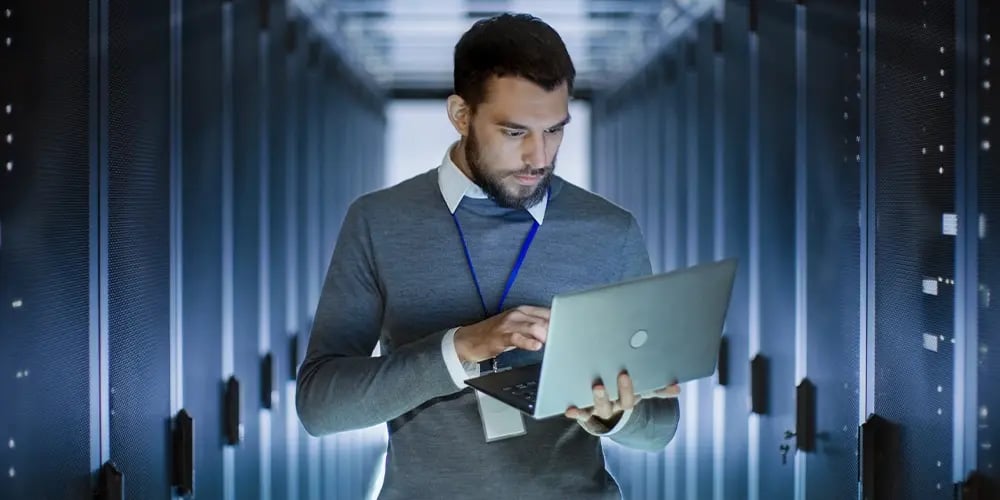 Person working on a laptop in a server room, focusing on data management and secure erasure processes.