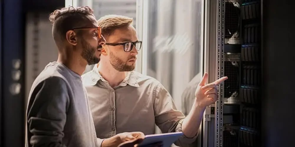 Two men in a server room, one pointing at a server rack, the other holding a tablet. They appear focused, suggesting a troubleshooting discussion.
