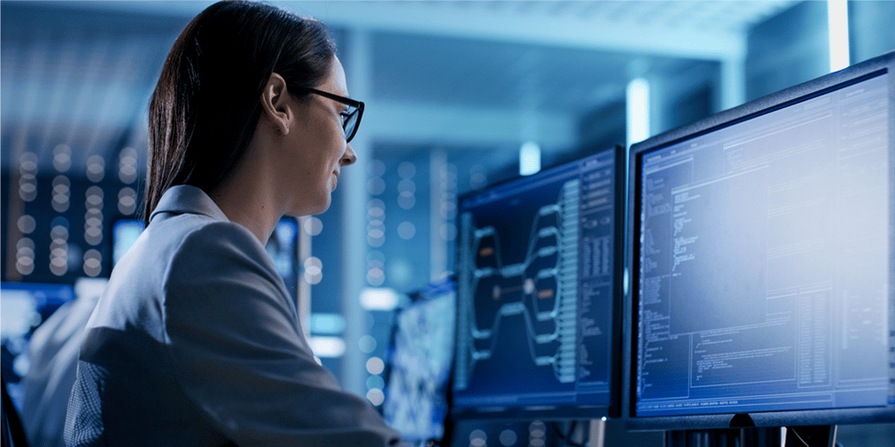 A woman in glasses and a blue blazer works at computer monitors displaying complex data and graphs in a dimly lit, modern office. The scene conveys focus.