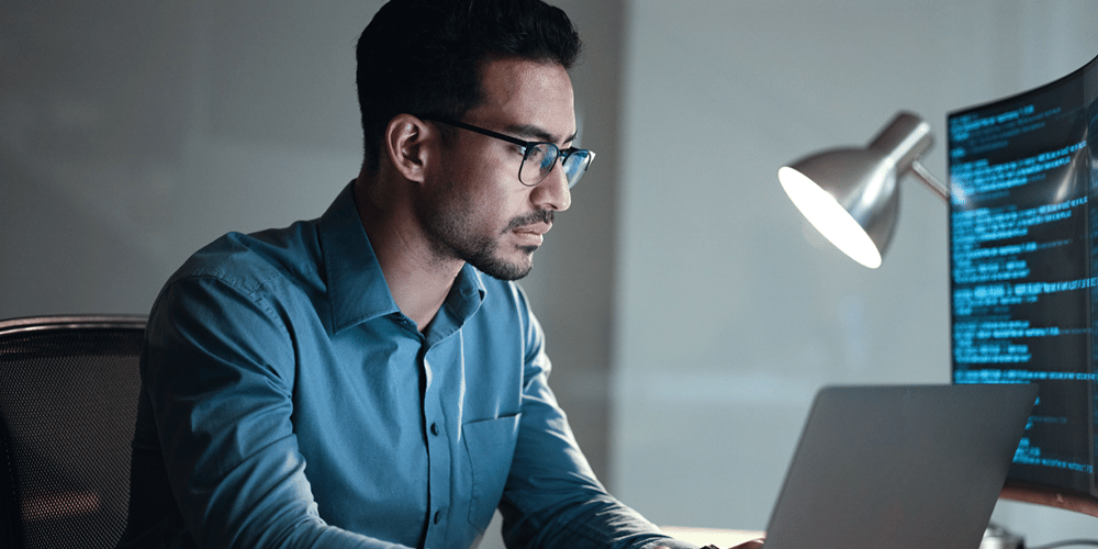 Man in glasses and blue shirt working on a laptop, with focus and concentration. A large monitor displays code. The scene is lit by a desk lamp.