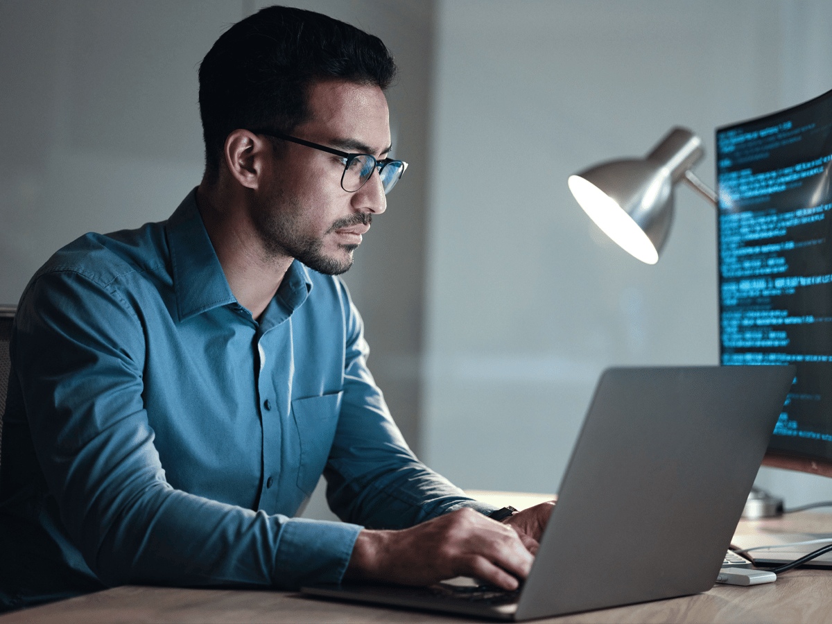 Man in glasses and blue shirt working on a laptop, with focus and concentration. A large monitor displays code. The scene is lit by a desk lamp.