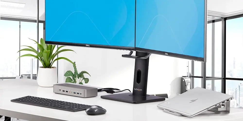 A clean workstation featuring dual Dell monitors, a StarTech docking station, keyboard, mouse, and laptop stand.