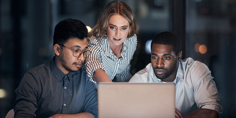 Three colleagues focus intently on a laptop in a dimly lit office, suggesting dedication and teamwork while solving a problem or project.