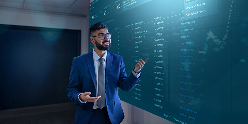 A man in a suit, smiling and gesturing toward a large digital screen displaying data and graphs. The setting is professional and tech-oriented.