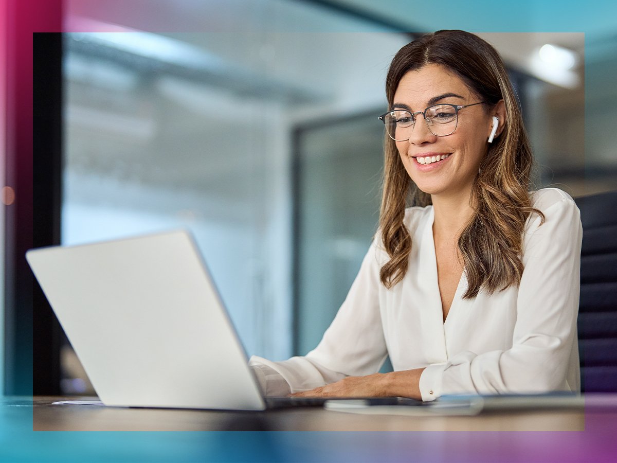 Person wearing glasses and wireless earbuds working on a laptop at a desk in a modern office.